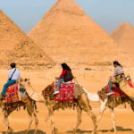 a group of travelers riding camels in front of the three pyramids 1200x5401828432573